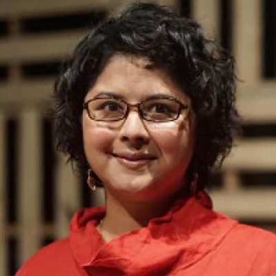 Nisha Sajnani wears an orange collared shirt, has black curly hair and square glasses