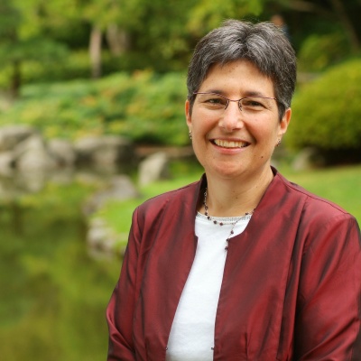 Tal Jarus wears a white t-shirt and red coat. They have salt-and-pepper hair and square glasses.